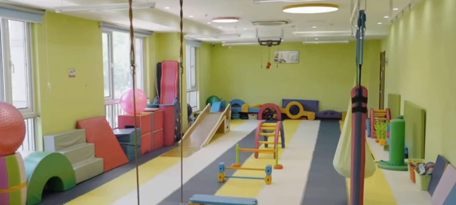 运动与感觉统合训练厅,天花板轨道上挂着悬吊设备,软垫地面上摆着滑梯、平衡木与训练器材