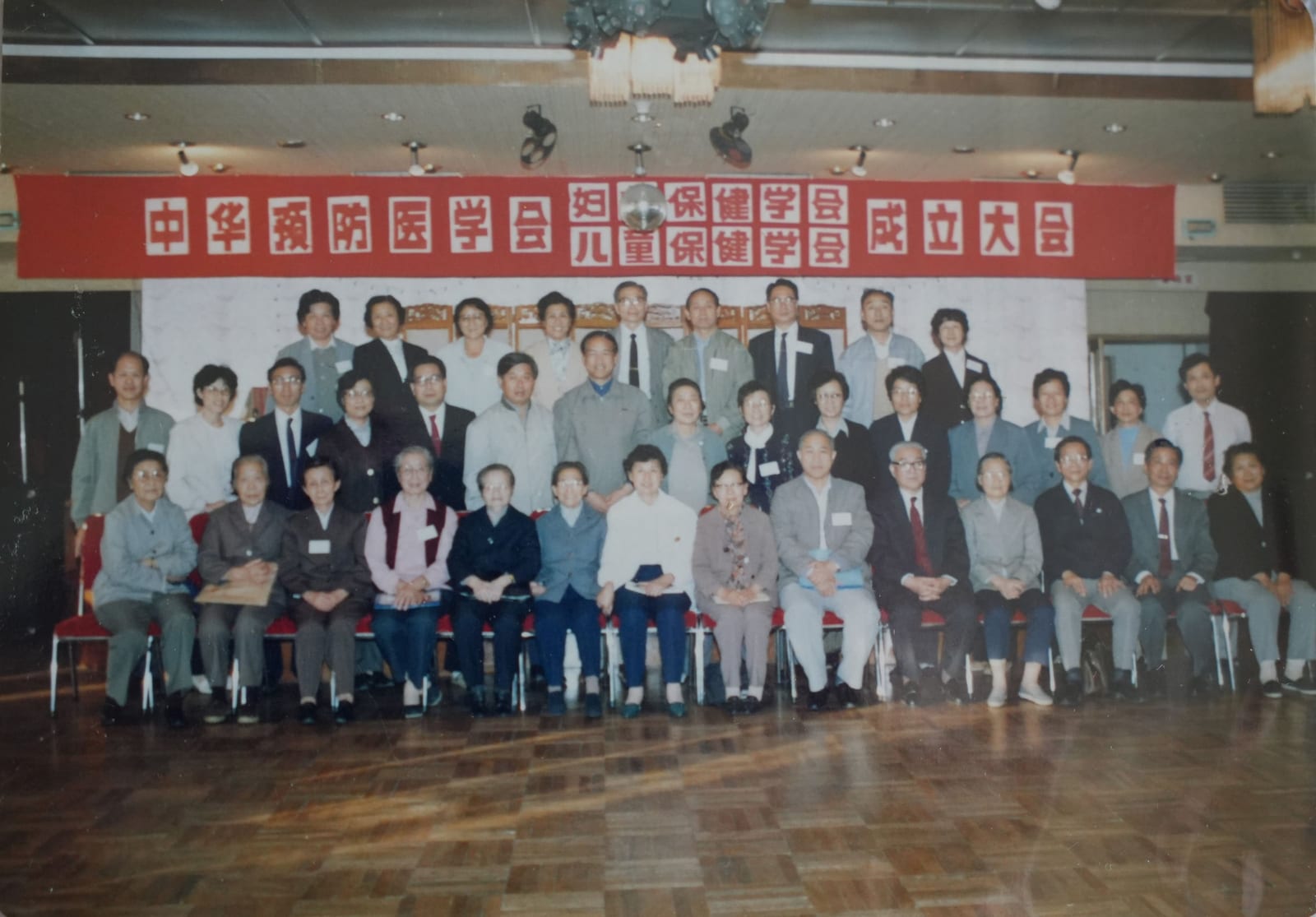 数十人分四排在会场里合影，身后横幅写着「中华预防医学会妇女保健学会、儿童保健学会成立大会」
