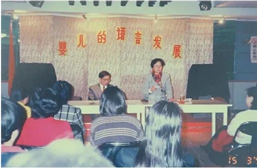鲍秀兰教授站在讲台后授课，身后幕布上贴着「婴儿的语言发展」，前排听众背对镜头