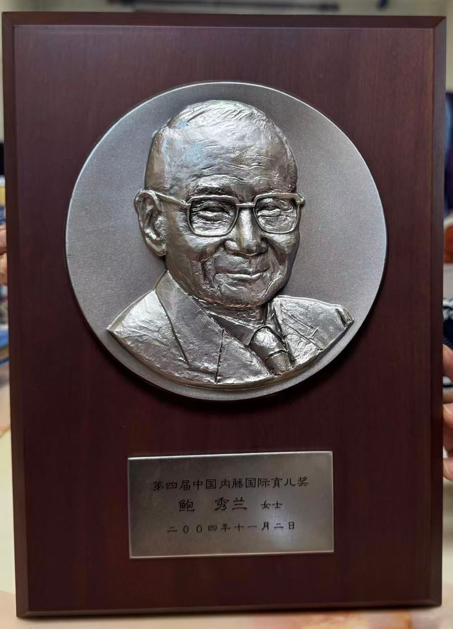 第四届中国内藤国际育儿奖奖牌：深色木底板上嵌着浮雕人像银牌，下方铭牌刻有「第四届中国内藤国际育儿奖　鲍秀兰女士　二〇〇四年十一月二日」