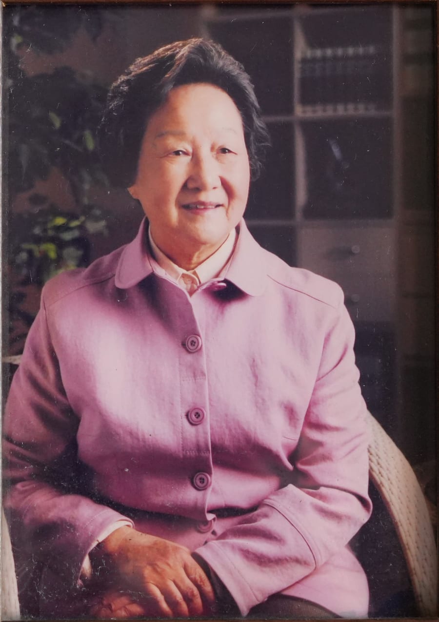 鲍秀兰教授的旧照：身穿粉紫色外套，坐在藤椅上微笑