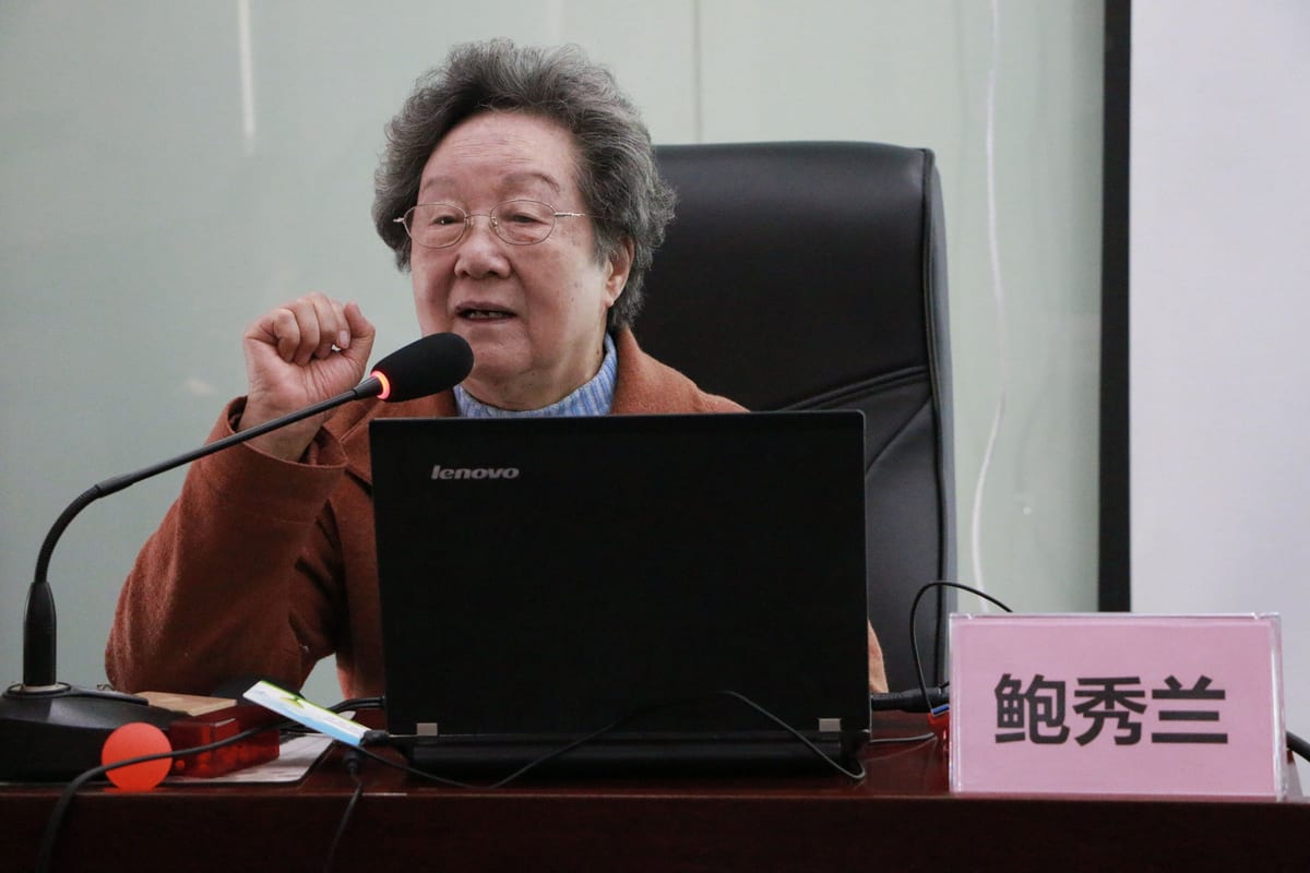 鲍秀兰教授坐在讲台后对着话筒讲课，面前是笔记本电脑与写着「鲍秀兰」的桌牌