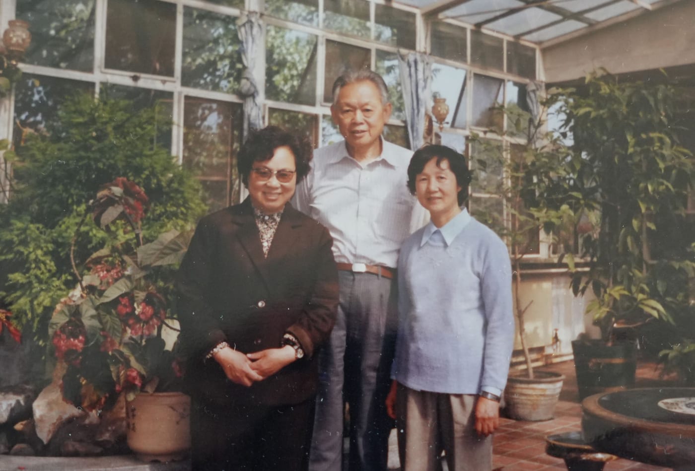 三人站在种满花木的温室里合影，中间是一位穿浅色衬衫的老先生