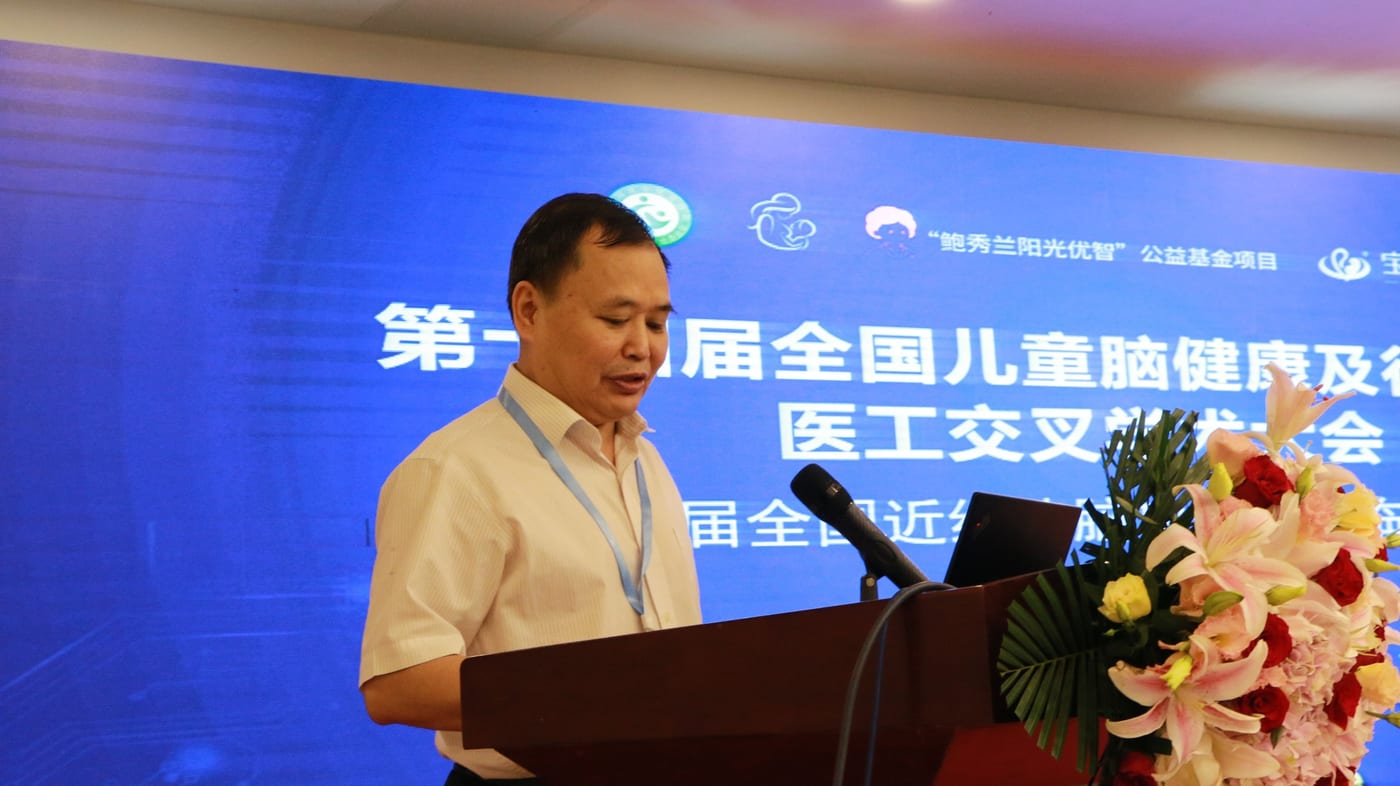 一位嘉宾身穿浅色条纹衬衫、挂着蓝色挂绳,在讲台后看着讲稿致辞,身后大屏写着「第十四届全国儿童脑健康及行为发育医工交叉学术大会」,讲台边摆着百合花束