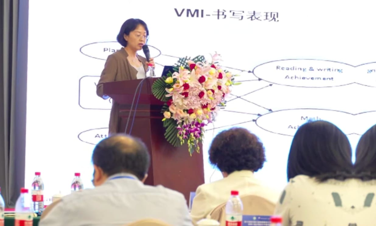 钱秋谨教授手持话筒在讲台后作报告,幻灯片标题为「VMI—书写表现」