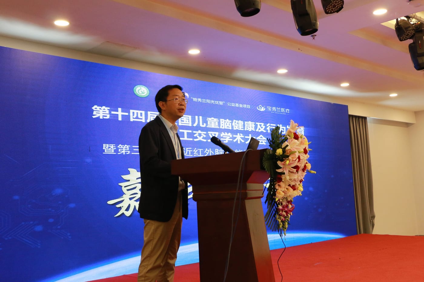 秦绍正教授身穿深色西装站在讲台前致辞,身后是大会背景板