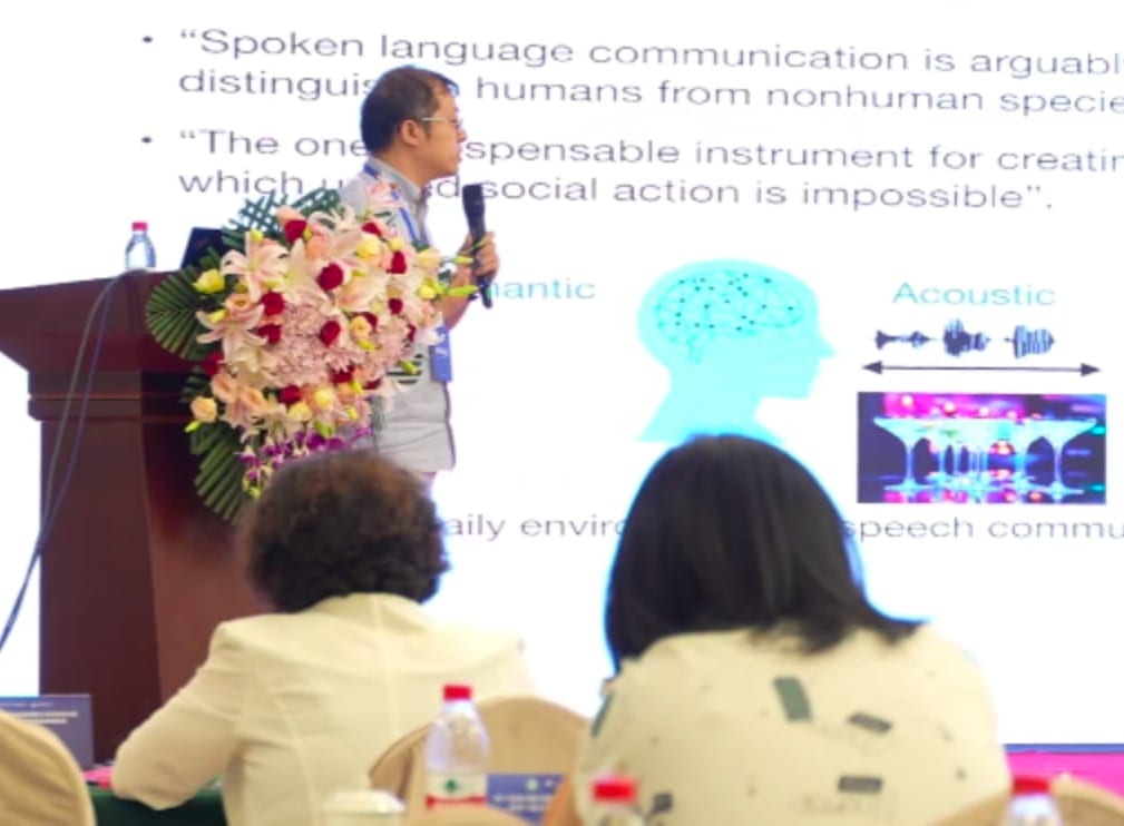 张丹教授手持话筒站在讲台旁作报告,幻灯片是关于口语交流的英文引文