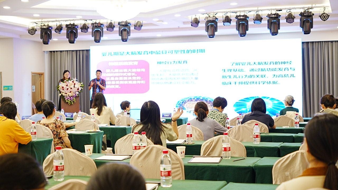 两人在舞台讲台旁作报告,大屏幻灯片讲婴儿期大脑发育的可塑时期,会场坐满听众