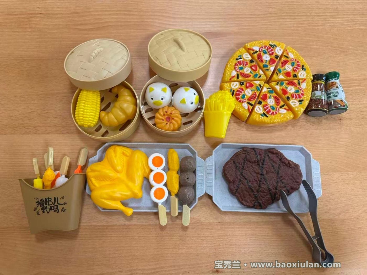 浅木色桌面上摆开的仿真食物玩具:两屉竹编蒸笼里装着玉米、牛角包与卡通造型的包子,两只灰色托盘上是整只烤鸡、鸡蛋、香肠、丸子串与一块牛排并配一把夹子,右侧是一块切成六块的比萨、一包薯条与两只调料瓶,左下角是一只装着串类的纸筒