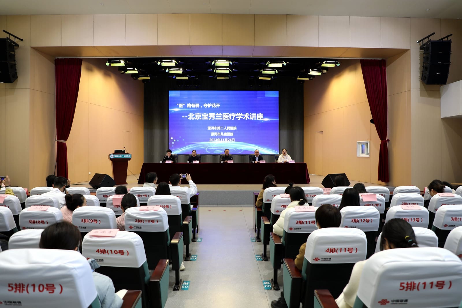 会堂里坐满听众,舞台大屏写着「"医"路有爱,守护花开——北京宝秀兰医疗学术讲座 漯河市第二人民医院 漯河市儿童医院 2024年11月24日」