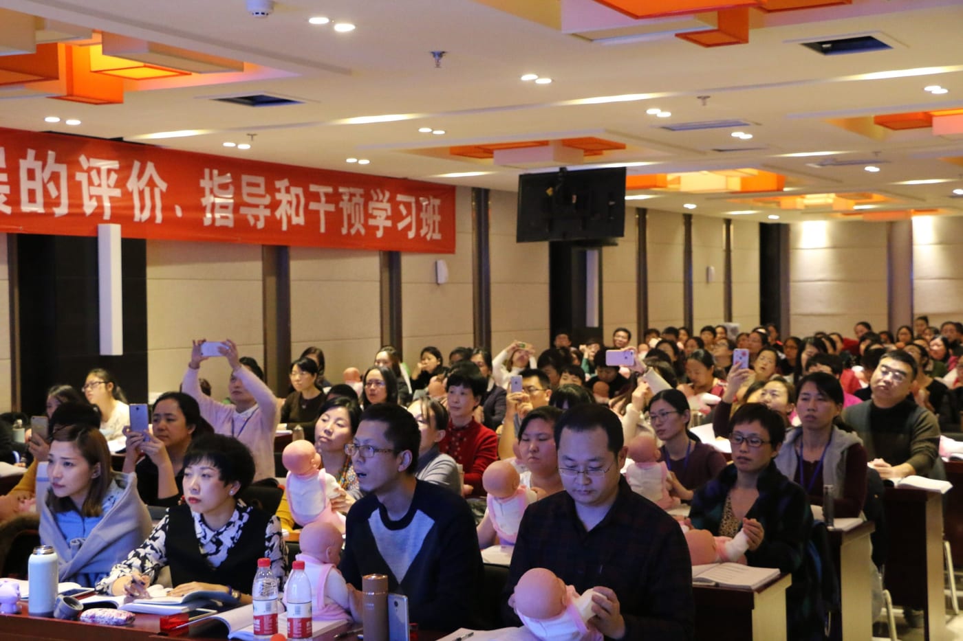学员坐满会场,怀里抱着婴儿模型练习,墙上横幅写着「……的评价、指导和干预学习班」