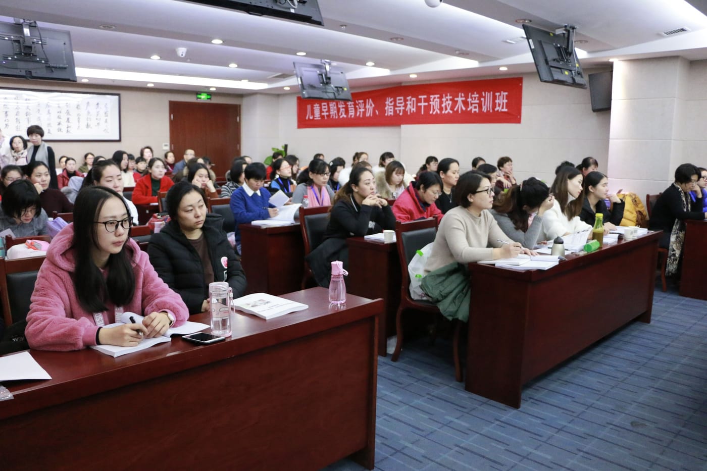 学员坐在课桌后听课记笔记,前方墙上挂着红色横幅「儿童早期发育评价、指导和干预技术培训班」