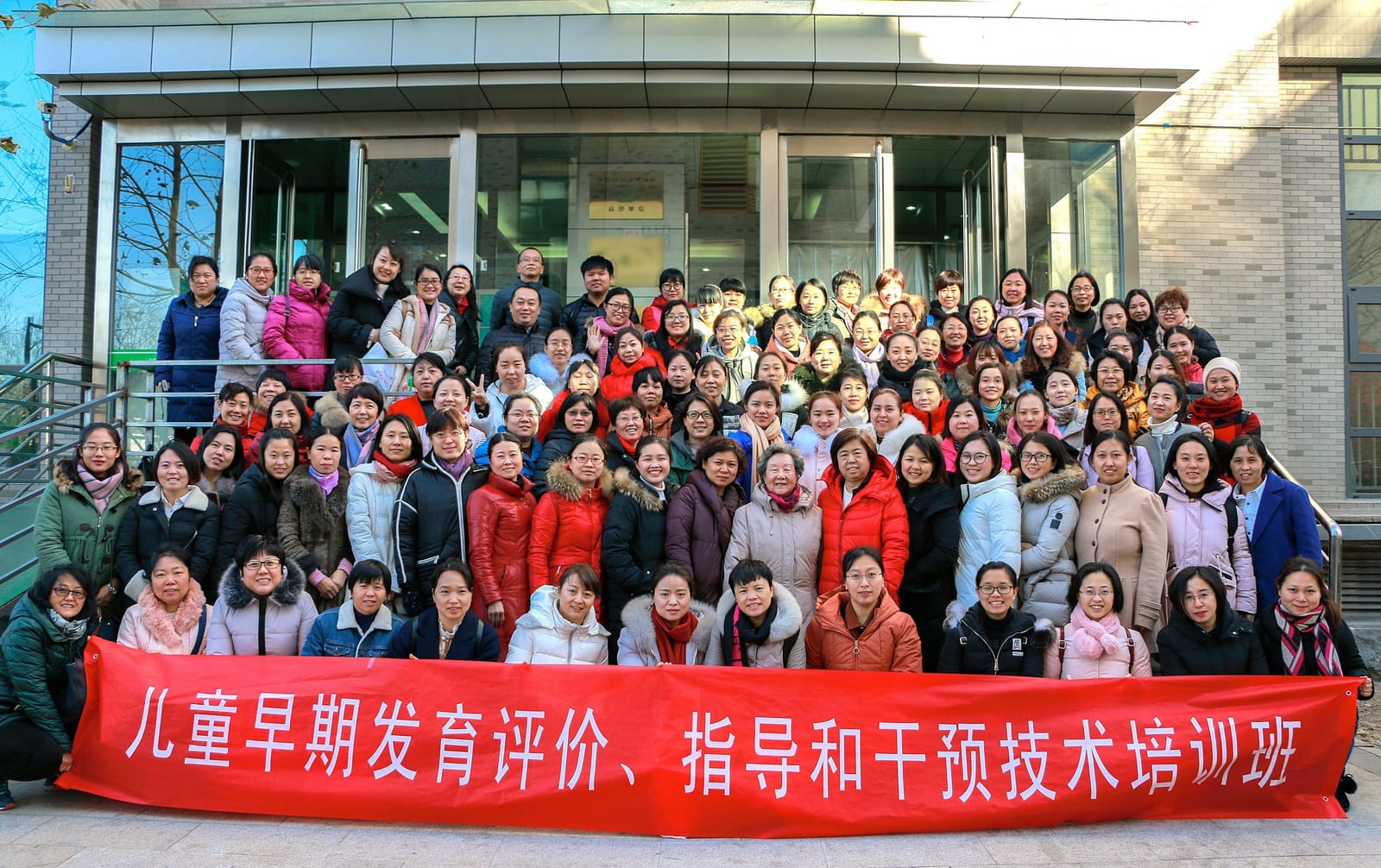 上百名学员在楼前台阶上合影,前排拉着红色横幅「儿童早期发育评价、指导和干预技术培训班」