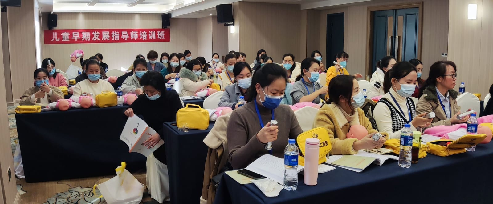 戴口罩的学员坐在长桌后看讲义,桌上放着黄色工具包与婴儿模型,墙上横幅写着「儿童早期发展指导师培训班」