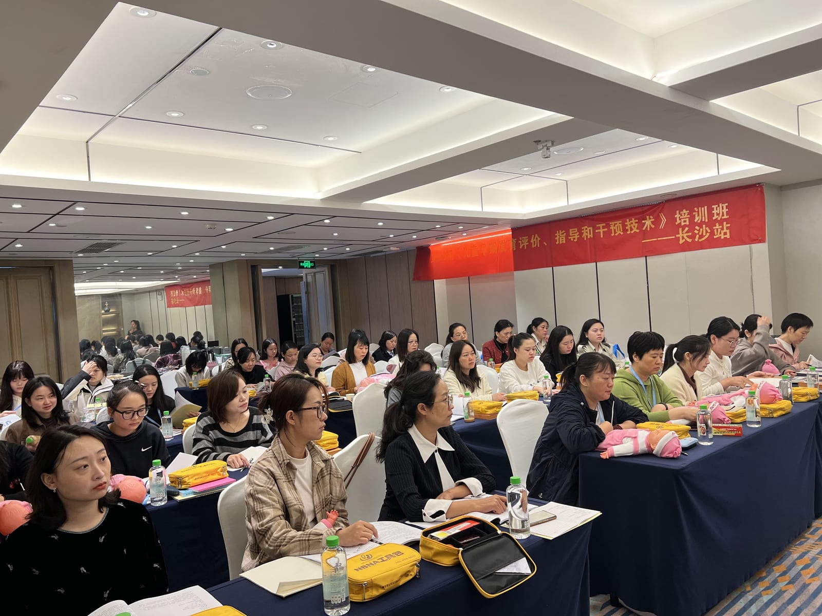 会议厅里坐满学员,桌上放着婴儿模型与黄色工具包,墙上横幅写着「《儿童早期发育评价、指导和干预技术》培训班——长沙站」