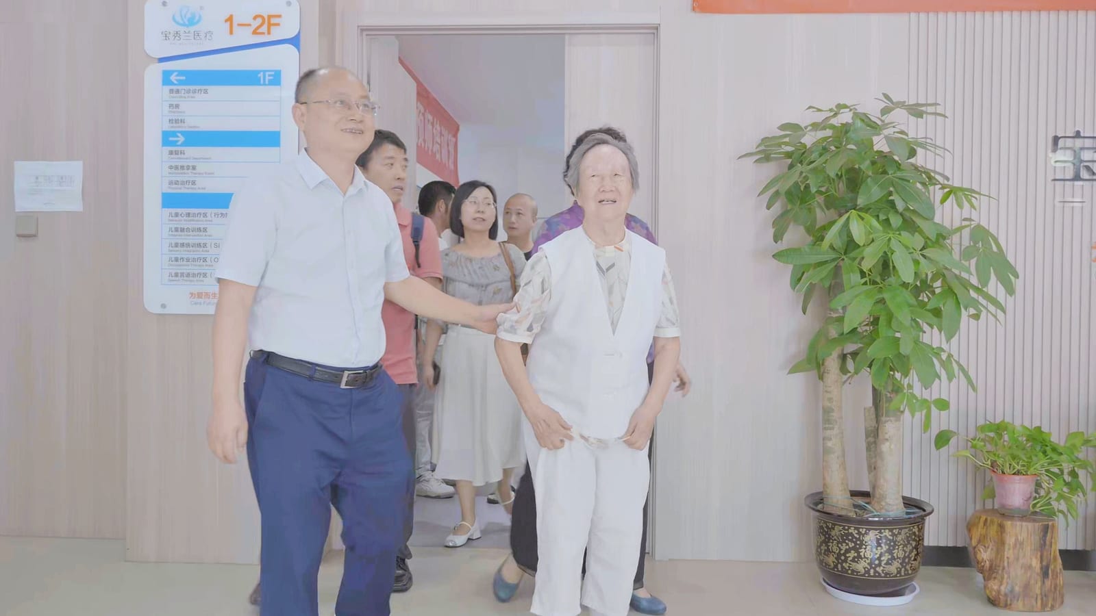 鲍秀兰教授与一位男士并肩走在门诊部走廊里,身后跟着几位来访者,墙上挂着「宝秀兰医疗 1–2F」的楼层指示牌