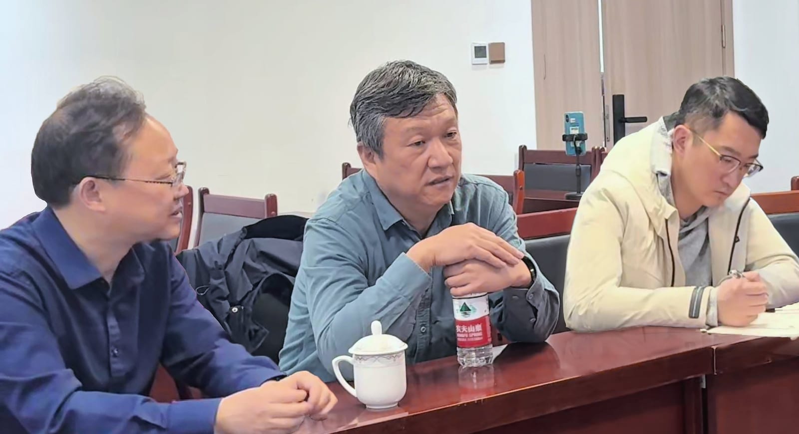 三位男士坐在会议桌旁交谈,桌上放着茶杯与矿泉水