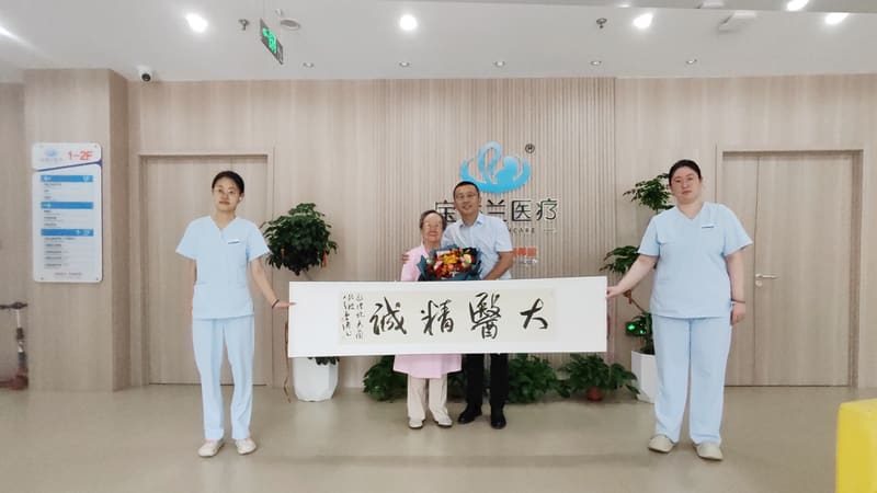 门诊大厅内，一位老人与一位医生手捧鲜花站在中间，两侧的护士与他们一起展开写着「大醫精誠」的锦旗